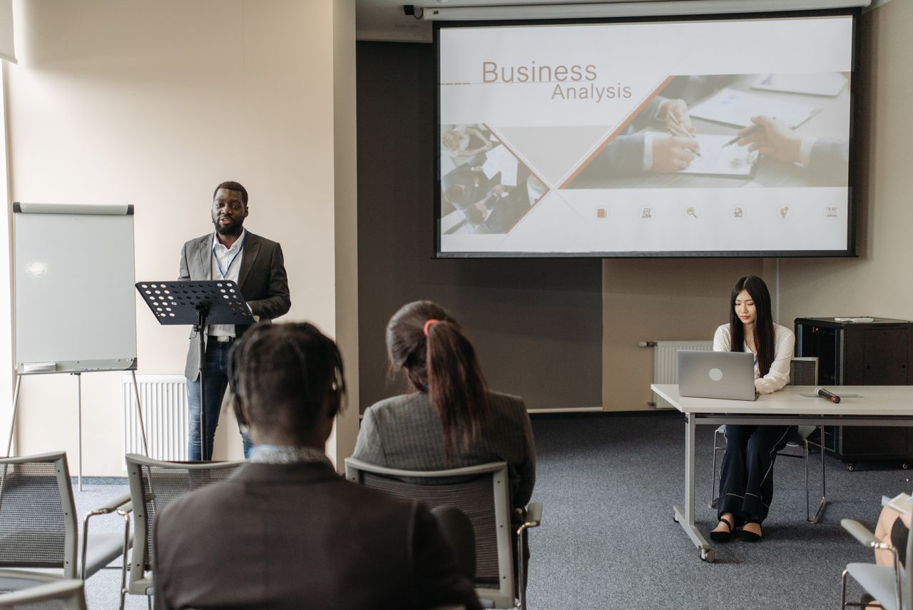 who-we-are Business seminar with a speaker and audience in a conference room.